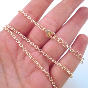 May include: A gold chain necklace with a lobster clasp closure. The chain is shown on a hand, with the chain laying flat.