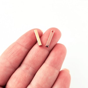 May include: Two gold-toned bar earrings, each with a small hole at the top. The earrings are held in a hand, with the background being a white surface.