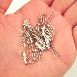 May include: A pile of silver-toned metal swivel clasps, also known as trigger snaps, in a hand. These clasps are used for attaching straps, cords, or chains to bags, clothing, or other items.