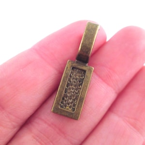 Puede incluir: Un colgante de metal dorado con forma rectangular y superficie texturizada. El colgante tiene un bucle en la parte superior para sujetarlo a una cadena o cordón.