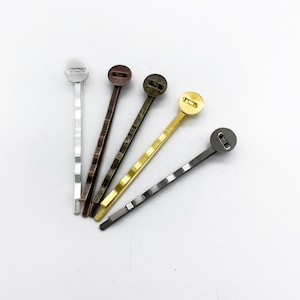 May include: Five hair clips with different colored metal tops. The clips are silver, bronze, gold, and two shades of gray.