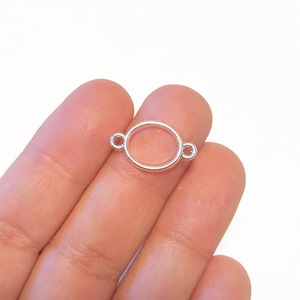 May include: A small, silver-colored oval charm with two loops on either side. The charm is held in a person's hand against a white background. The charm is likely used for jewelry making or crafting.