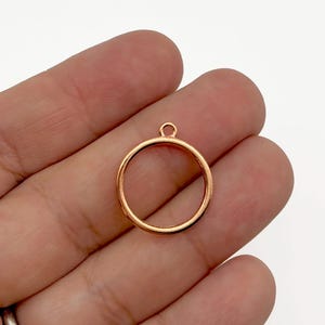 May include: A rose gold-colored metal circle pendant with a small loop at the top. The pendant is held in a person's hand. The pendant is a jewelry finding, suitable for making necklaces, earrings, or other accessories.