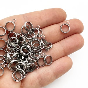 May include: A pile of silver-toned metal jump rings with a loop on one side and a small loop on the other side. The jump rings are in a hand.