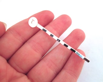 Può includere: Un fermaglio per capelli in argento con una piccola parte superiore rotonda che presenta la lettera "T" incisa su di essa. Il fermaglio ha un design rigato.