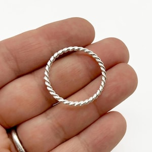 May include: A silver-colored ring with a twisted rope design. The ring is held in a hand against a white background. The ring has a simple, elegant design.
