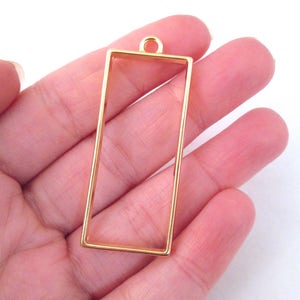 Pode incluir: Pingente de moldura de metal dourado com formato retangular e um laço na parte superior para pendurar.