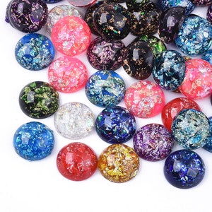 Könnte beinhalten: Sortiment runder, irisierender Glascabochons mit goldenen und silbernen Flocken. Die Cabochons sind in verschiedenen Farben, darunter Blau, Grün, Rot, Rosa und Lila.