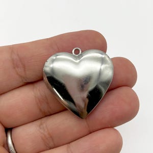 May include: A shiny, silver heart-shaped pendant with a small loop at the top for hanging. The pendant has a smooth, reflective surface, and the heart shape is clearly defined. The overall design is simple and elegant.