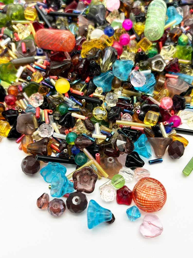 May include: A large assortment of colorful glass beads in various shapes and sizes, including round, square, star, and flower shapes. The beads are scattered on a white surface.