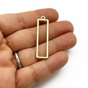 Puede incluir: Un colgante rectangular de color dorado con un pequeño lazo en la parte superior. El colgante se sostiene en la mano de una persona sobre un fondo blanco. El colgante es un diseño simple de marco abierto, adecuado para la fabricación de joyas.