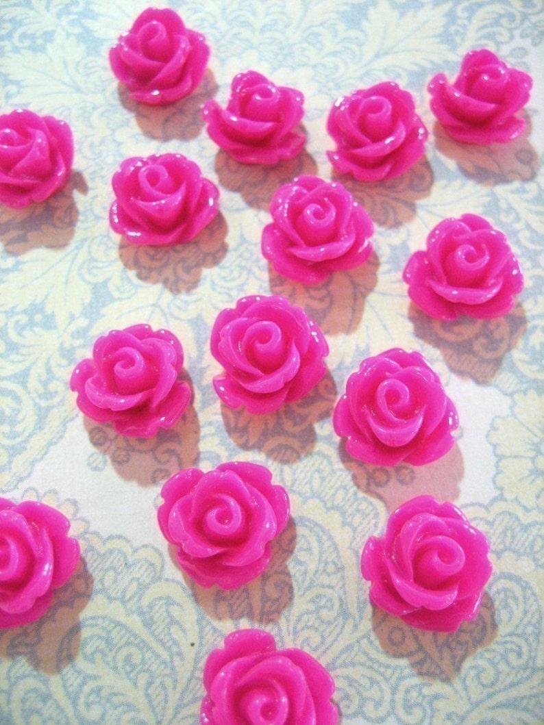 May include: A collection of 14 bright pink plastic rose embellishments. The roses are all the same size and shape, and they have a glossy finish.