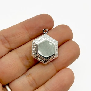 May include: A silver-colored, hexagonal locket with an ornate, scrollwork design around the edges. The locket has a small loop at the top for attaching to a chain. The locket is held in a hand.