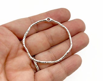 Könnte beinhalten: Ein silberfarbener, strukturierter Metallreifen mit einem kleinen Loch oben. Der Reifen ist kreisförmig mit einem unebenen, gehämmerten Aussehen. Das Schmuckteil wird in einer Hand vor weißem Hintergrund gehalten.