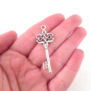 May include: Silver key charm with an ornate design featuring two hearts. The key has a loop at the top for attaching to a necklace or bracelet.