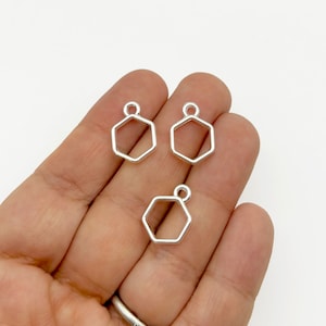 May include: Three silver hexagon shaped charms with a loop at the top. The charms are on a hand.