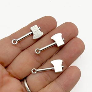 May include: Three small, silver-colored axe charms. Each charm features a detailed axe head and a loop for attaching to jewelry or other crafts. The axes are held in a hand against a white background.