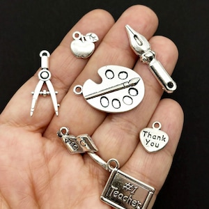 May include: A collection of silver-toned metal charms featuring school-themed designs including an apple, a drafting compass, a paint palette with a brush, a fountain pen, an open book with ABC letters, a heart with Thank You, and a chalkboard sign.