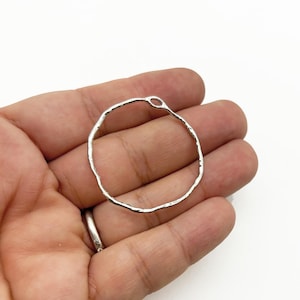 May include: A silver-colored, hammered metal hoop earring with an irregular, textured surface. The earring has a small loop at the top for attachment. The earring is held in a hand against a white background.