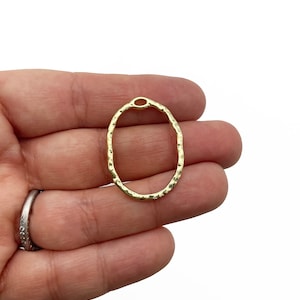 May include: Gold oval-shaped metal pendant with a hammered texture. The pendant has a small hole at the top for attaching a chain or cord.