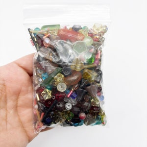 May include: A clear plastic bag filled with a variety of colorful glass beads. The beads are in various shapes and sizes, including round, square, and star-shaped. The beads are a mix of red, blue, green, yellow, and clear.