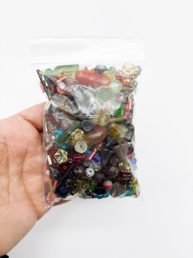 May include: A clear plastic bag filled with a variety of colorful glass beads. The beads are in various shapes and sizes, including round, square, and star-shaped. The beads are a mix of red, blue, green, yellow, and clear.