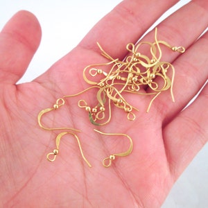 May include: A collection of gold-colored earring hooks, some with small ball accents. The hooks are curved and designed for jewelry making. The hooks are held in a person's hand against a white background.