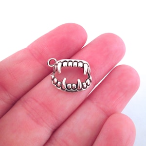 May include: Silver charm in the shape of a set of vampire fangs. The charm is detailed with small, round teeth and a pointed canine tooth on each side.