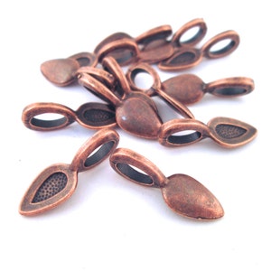 Op de afbeelding: Een groep van 10 koperkleurige metalen hangerbedels met een traanvorm en een kleine lus aan de bovenkant. De bedels liggen verspreid op een wit oppervlak.