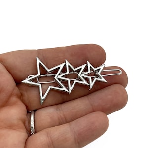 May include: Silver hair clip with three star-shaped cutouts in a row. The stars are linked and attached to a barrette clip. The clip is made of a shiny, metallic material.