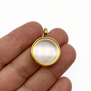 Peut inclure: Un pendentif de médaillon rond de couleur or avec un devant en verre ou en plastique transparent. Le pendentif a une petite boucle en haut pour une chaîne. Le médaillon est tenu dans la main d'une personne sur fond blanc.
