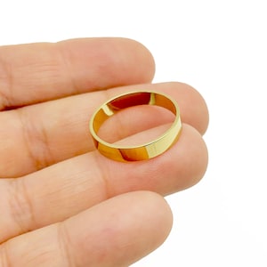 May include: A gold-colored band ring with a flat, polished surface. The ring is held in a hand against a white background. The ring appears to be a simple, classic design, suitable for everyday wear or as a wedding band.