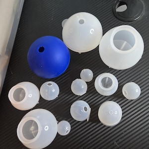 May include: A collection of spherical silicone molds in various sizes, including one solid blue sphere mold and several translucent white sphere molds. Each mold features a small circular opening at the top, designed for casting or crafting purposes.