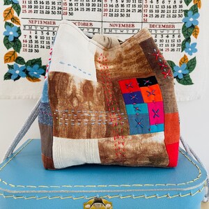 May include: A patchwork fabric bag with a drawstring closure, featuring a mix of brown, white, blue, and red fabrics. The bag is embellished with colorful cross-stitch details and sits atop a blue suitcase.