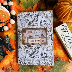 May include: A Halloween-themed journal with a black and white floral and skull pattern. The journal features a rectangular label with the words "Hocus Pocus Apothecary". A black cat figurine, a pumpkin decoration, and a "Trick or Treat" sign are also visible.