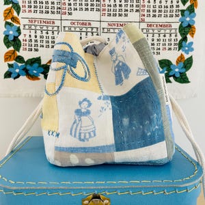 Pode incluir: Uma bolsa com cordão patchwork com um cordão creme, com painéis de tecido azul e amarelo com ilustrações em estilo vintage. A bolsa está sobre uma mala azul, com um calendário ao fundo.