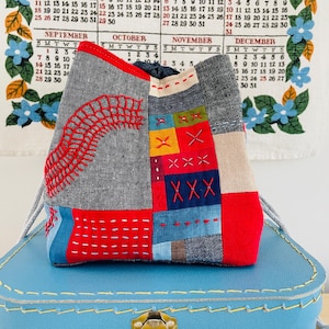 Op de afbeelding: Een patchwork tas met een trekkoordsluiting, met een mix van grijze, rode, blauwe en beige stoffen. De tas is versierd met rode geborduurde details en kleurrijke gestikte vierkanten. Hij rust op een blauwe koffer.