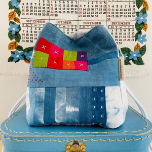 Könnte beinhalten: Eine handgefertigte Denim-Tasche mit einem Patchwork-Design mit roten, rosa, grünen und orangefarbenen Quadraten. Die Tasche hat einen weißen Kordelzug und steht auf einem blauen Koffer. Das Etikett lautet "KMS Handmade".