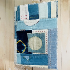 May include: A rectangular fabric journal cover featuring a patchwork design of various blue and white textile scraps. The surface includes visible hand-stitched embroidery details, including red and yellow thread accents, and a central circular patch.