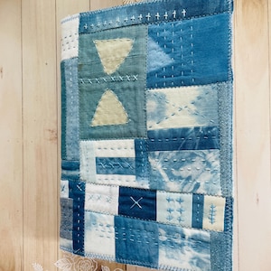 May include: A rectangular fabric cover featuring a patchwork design of various blue and white textile squares. The surface is decorated with visible white hand-stitched embroidery patterns, including crosses, lines, and two light-colored triangles.