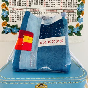 Può includere: Una borsa in tessuto blu con chiusura a coulisse caratterizzata da un design patchwork con un quadrato rosso e giallo, punti a croce ricamati in bianco e un pannello blu scuro con macchie bianche. La borsa ha cordini bianchi e dettagli cuciti a mano.