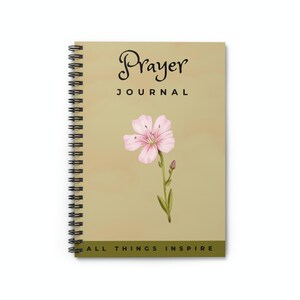 以下が含まれることがあります： ベージュのスパイラルバインダーノート。表紙には黒い筆記体で「Prayer Journal」と書いてあります。ピンクの花と緑の葉が表紙の中央に配置されています。表紙の下部には「All Things Inspire」と書いてあります。