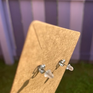 May include: A pair of silver stud earrings with clear plastic backs. The earrings are displayed on a brown card.