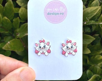 May include: A pair of stud earrings featuring a white cartoon cat with a pink bow on its head and a pink collar, displayed on a white card with the text you can fly design co printed in a circular lavender logo at the top center of the card.