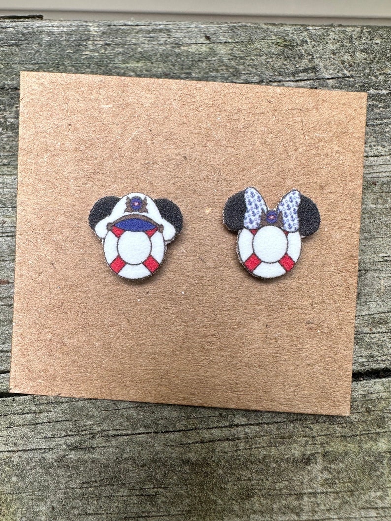 May include: A pair of wooden Mickey Mouse ears with a nautical theme. One ear features a red, white, and blue life preserver with a captain's hat on top. The other ear features a red, white, and blue life preserver with a blue and white bow on top.