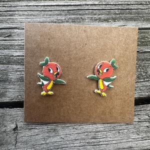 May include: A pair of orange bird earrings with green leaves on a brown card. The birds are cartoon-like and have yellow beaks and white eyes.