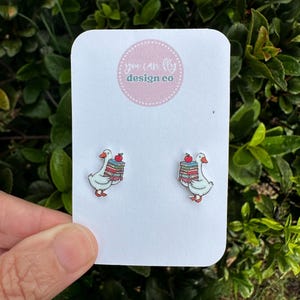 May include: A pair of white goose stud earrings featuring a stack of colorful books and a red apple on top. The earrings are displayed on a white rectangular card with the text you can fly design co printed in a circular pink logo at the top.