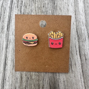 May include: A pair of stud earrings featuring a cartoon hamburger with a smiling face and a cartoon container of french fries with a smiling face, both displayed on a brown rectangular card with a circular hole at the top.