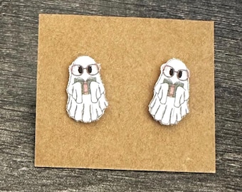 May include: A pair of white ghost earrings with black glasses and a pink book. The earrings are on a brown card.