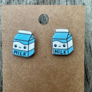 May include: A pair of milk carton earrings with a cute cartoon face and the word "MILK" printed on them. The earrings are on a brown card.